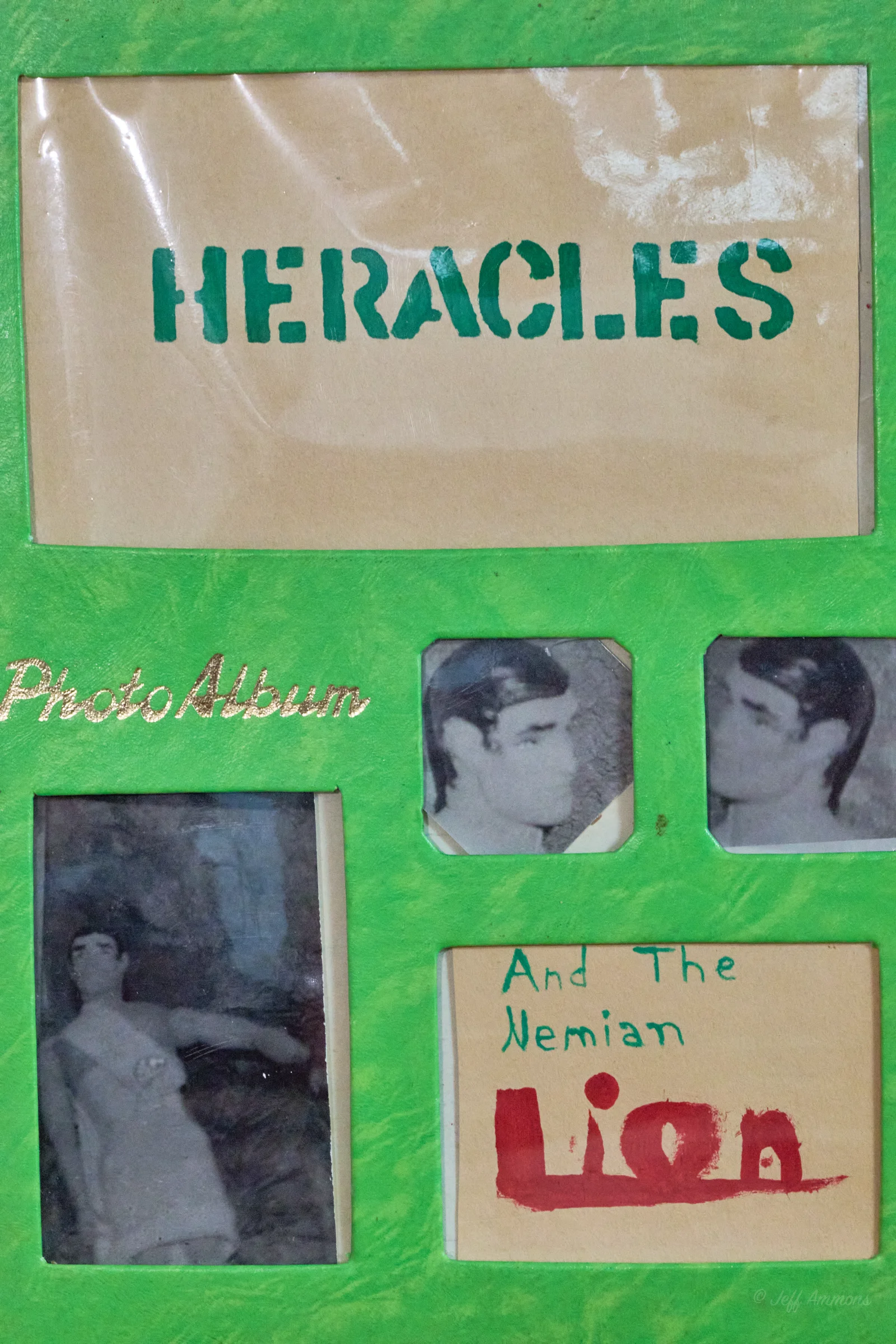 A green scrapbook cover reading 'HERACLES Photo Album' with several cut-out black-and-white photos of an action figure, and a hand-lettered caption reading 'And The Nemian Lion.'