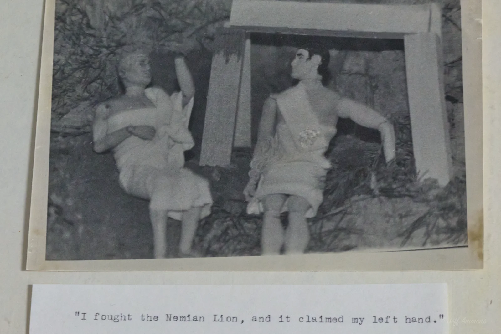 Heracles sits beside a companion figure with an arm wrapped in a sling, in front of the stone doorway.