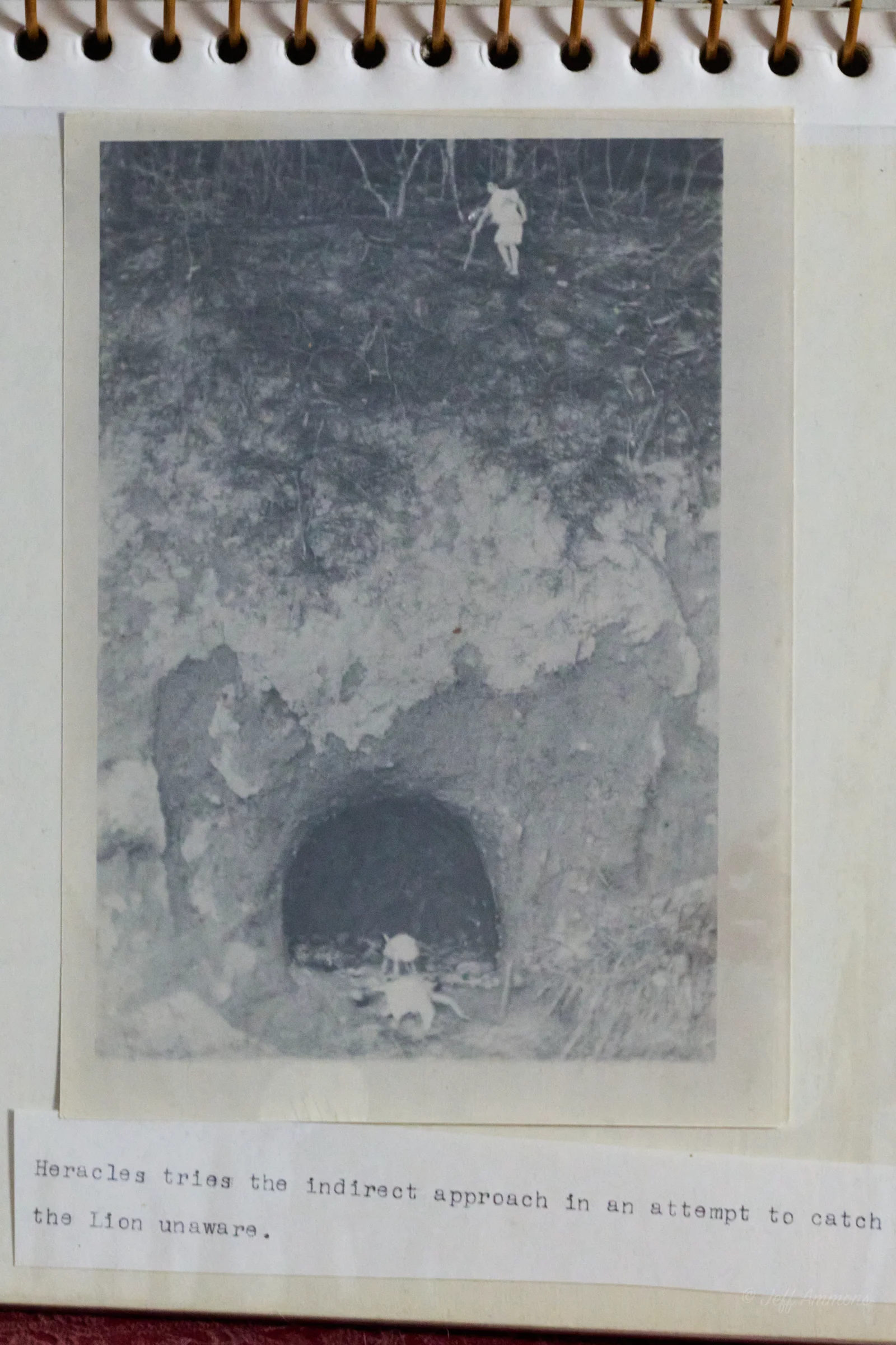 A tiny figure of Heracles approaches a dark cave entrance in a rocky hillside, seen from a distance above.